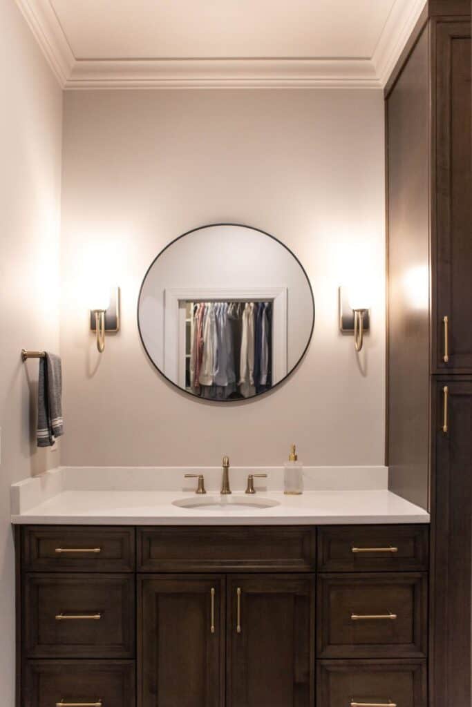 Elegant primary bathroom featuring dark wood cabinets, a round mirror, and wall-mounted lighting fix.