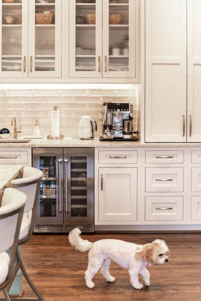 Bright white kitchen with glass-front cabinets, coffee station, and a small dog on hardwood floor.
