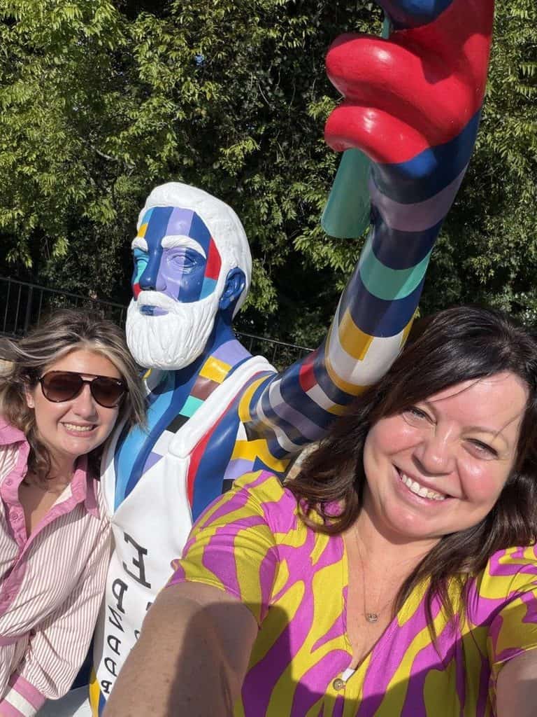 Two women taking a selfie with a vibrant, colorful sculpture of a bearded man.